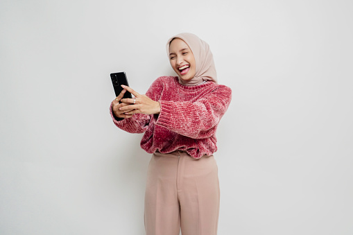 A portrait of a happy Asian woman wearing a pink sweater and hijab, holding her phone, isolated by white background