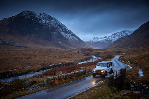 Land Rover Defender 4x4 on the road with lights on, Glen Etive, Scotland, UK during winter time, there is snow on the mountains an the road is leading into the distance. It’s raining, the sky is dark, the car is driving over a bridge.