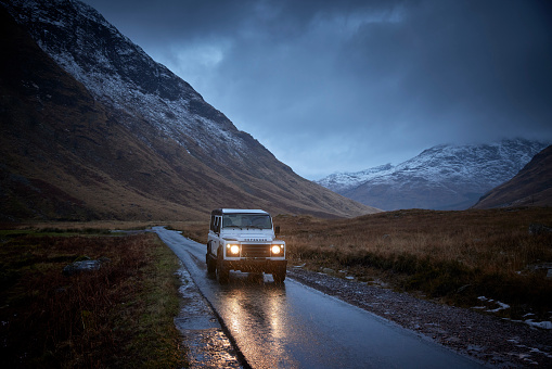 Land Rover Defender 4x4 on the road with lights on, Glen Etive, Scotland, UK during winter time, there is snow on the mountains an the road is leading into the distance. It’s raining, the sky is dark, the car is driving over a bridge.