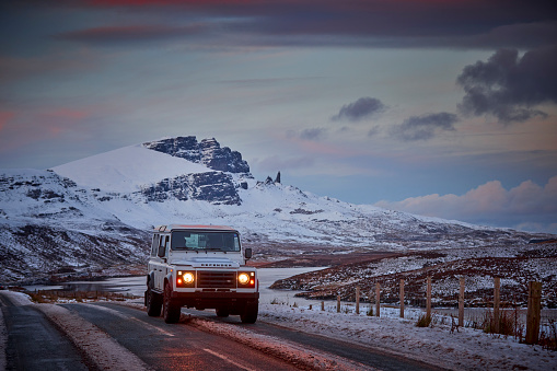 Landrover Defender 4x4 is driving on a road, in the back you see Old man of Storr, UK, Isle of Skye. There are electrical cables on the side of the road, and mountains in the background. Everything is covered with snow. It's getting dark
