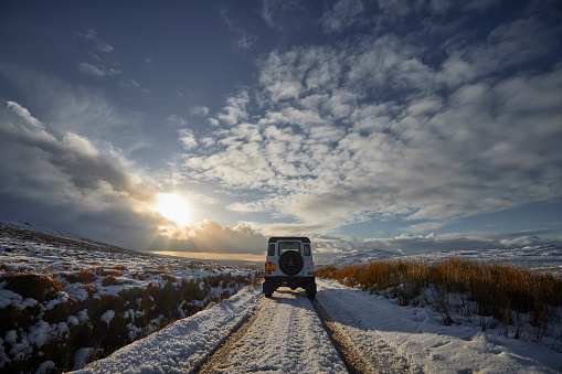 Landrover Defender 4x4 is driving on a snow covered road, through Scotland, UK, Isle of Skye. The sun is shining and the are some clouds in the sky. Everything is covered with snow.