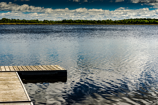 Wooden Dock on small lake covered in ripples with tree lined shore in the distance under a blue cloudy sky