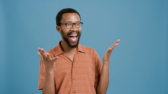 Excited, black man and surprise with hands out for good news or winning on a blue studio background. Face of happy African or male person with smile for deal, alert or sale promotion on mockup space
