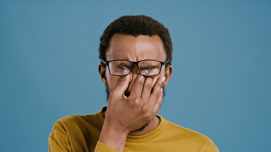 Man, glasses and headache in studio for stress with vertigo, eye strain or blurry vision from burnout. African person, migraine and brain fog with mental health crisis and anxiety on blue background