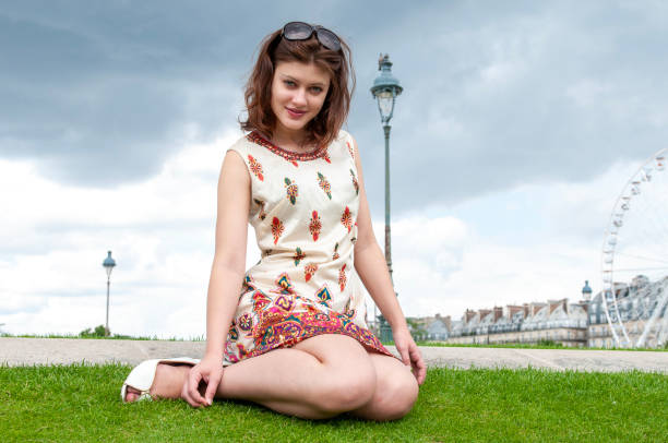 full length shot of an elegant young brunette in colourful dress wearing sunglasses in her hair sitting on the grass in le Jardin des Tuileries in Paris on a cloudy day looking at the camera
