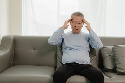An unhappy retired Asian man wearing a t-shirt has a headache while sitting on the sofa at home, has a stress headache, holds his hand to cover his forehead in pain.