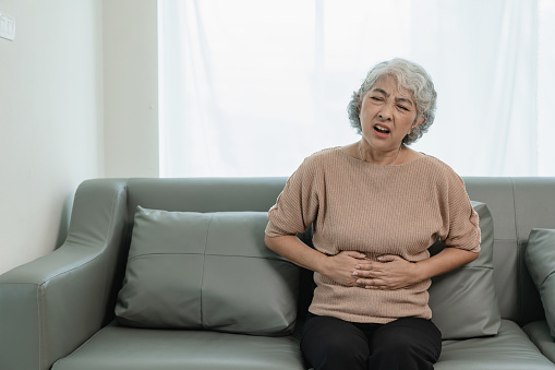 An elderly woman has severe abdominal pain. Sitting alone on the sofa while having dinner at home, elderly health care concept. Illness. Health insurance.