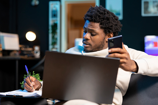 Self employed man working from home, writing down contact details after talking on phone with client. BIPOC man using pen to note information on notepad while teleworking in apartment