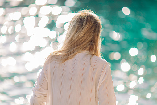 Young woman standing by the sea and looking at it. He hair is tousled by the breeze.