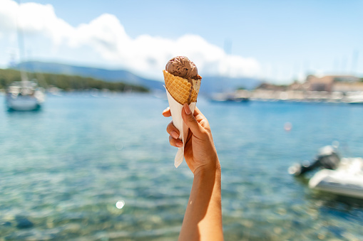 Hand is holding chocolate ice cream against bay with beautiful blue water and small marina. Fiscardo, Kefalonia island, Greece