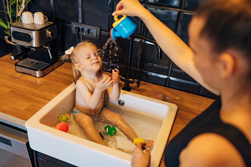 A young mother and her cute baby girl play with a watering can, while the girl bathes in the kitchen sink