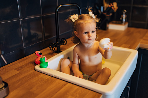 A baby girl is sitting in the kitchen sink, playing with toys, enjoying the fun