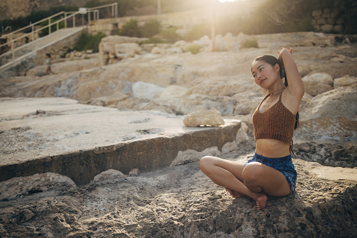 An Asian woman sits in the lotus position on a rocky seashore and does yoga and stretching exercises