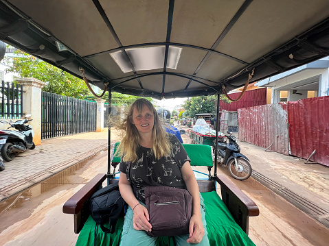 An Image of a mature lone female riding at the back of a Tuc-Tuc on a street in Cambodia. She is smiling at the camera. Either side of this lady there is a residential area, her hair is lifted by the speed of travel. an uplifting experience.