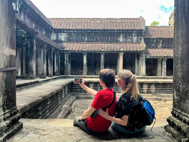 Mother and teenage son  are taking a selfie whilst sitting on a step looking at a view of a temple at Angkor Wat. This image is taken from the rear , a vacation moment. The lady is lovingly close to her child, she is wearing a blue backpack, whilst he is wearing a red tee shirt.