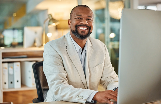 Portrait, business and happy black man on computer in office, workplace and company at night for career. Face, advisor and smile of entrepreneur, professional and African employee working in Kenya