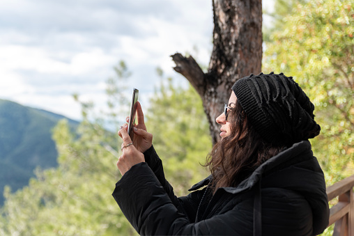 beautiful middle-aged woman wearing a woolen hat takes photos with her phone in nature