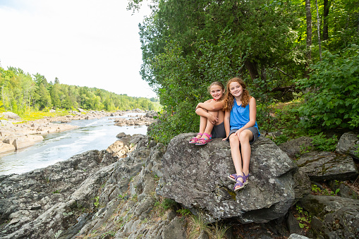 Two young girls (sisters) sitting on a large rock on the bank of the St. Louis River in Minnesota, USA. Both girls are smiling and looking at the camera.