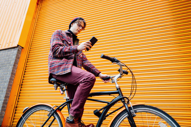 istock Young hipster man using mobile phone while sitting on cruiser bicycle near orange wall. Student using 5g network,rests after riding. Eco friendly transport for carbon footprint. Sustainable lifestyle 2043317211