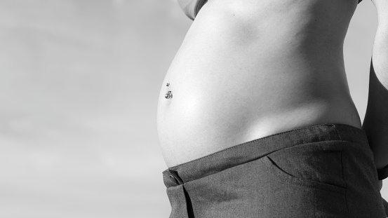 Close-up view of pregnant belly outdoors in black and white with navel piercing in the first months of pregnancy. Side view of pregnant belly with sky in the background and  copy space