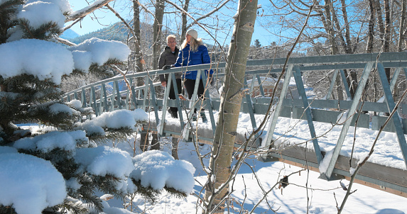 Mature couple cross bridge in forest after fresh snow fall, mountains distant