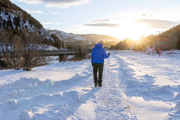 Mature man walks along pathway and uses mobile phone on a cold day as sun rises over distant mountains