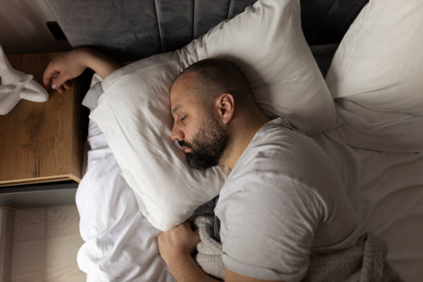 Bed, morning and relax man with beard sleeping, tired or nap for relief, wellness and rest in home, house or apartment. Fatigue, comfort and face of person cozy, dream and exhausted on bedroom pillow stock photo vista superior dormitorio del apartamento: joven guapo durmiendo cómodamente en una cama en su dormitorio por la mañana. cómodo apartamento con chico descansando para el productivo día siguiente. de arriba hacia abajo arriba foto de archivo