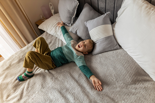 Portrait of little, smiling caucasian boy on the bed at home. Cute child relaxing, resting in bedroom. Positive emotions. Cozy and modern interior. Natural, earth colors. Casual clothes. stock photo