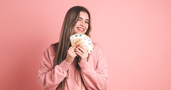 Happy young female model in pink hoodie with long blond hair looking at camera while holding fanned out 50 euro banknotes over pink background