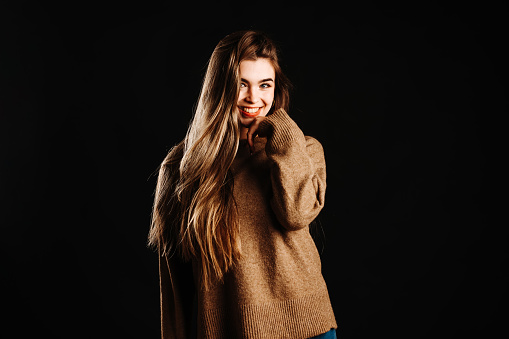 Portrait of happy young female model in brown sweater with long blond hair looking at camera while standing with hand at chin against black background