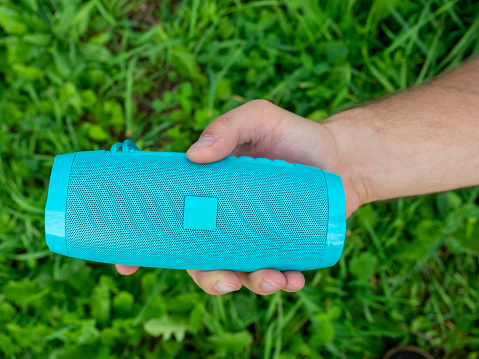Close-up of a man's hand holding a blue wireless music speaker. There is a thick green grass in the background. Using an outdoor device. Top view