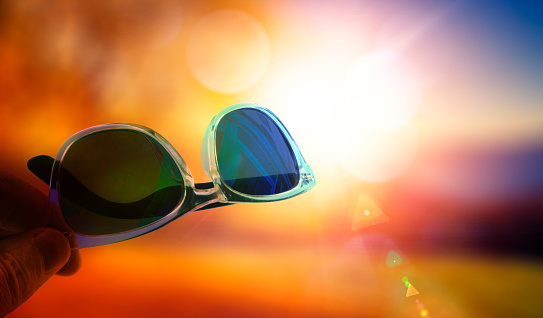 Hand holding sunglasses on a beach at sunset.