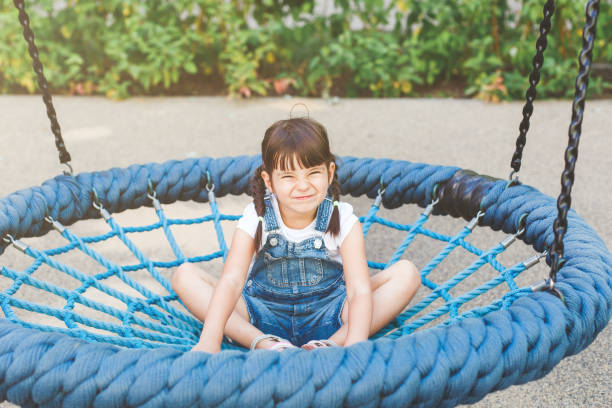 one little cute girl 4 years old sits on a hanging round swing with a net and smiles funny on the playground in summer, childhood concept. - child 4 5 years laughing little girls - fotografias e filmes do acervo