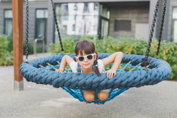one little cute girl 4 years old sits on a hanging round swing with a net and smiles funny on the playground in summer, childhood concept. - child 4 5 years laughing little girls - fotografias e filmes do acervo
