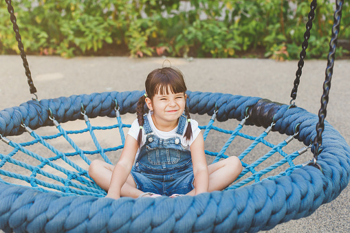 one little cute girl 4 years old sits on a hanging round swing with a net and smiles funny on the playground in summer, childhood concept