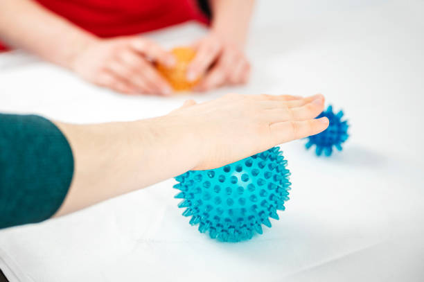 A woman rolls her hand on a hedgehog ball under the supervision of a medical staff member during a physiotherapy treatment.