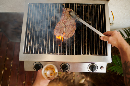 Man hand, cooking food or meat on barbecue grill, fire or grid for lunch or dinner at house. Hands of person outdoor in backyard with a grilled beef steak with smoke and tongs pov at protein bbq