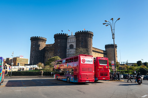 Naples, Italy - December, 18, 2022: Majestic medieval Castel Nuovo with a double-decker tour bus parked, tourists milling about in the foreground
