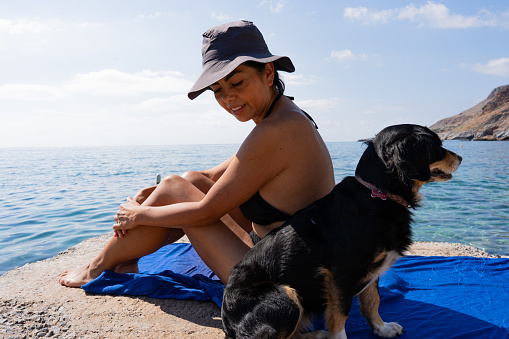 Latin woman sitting on the pier enjoying a sunny morning on the Greek island of Crete with her pet and with the Aegean sea in the background