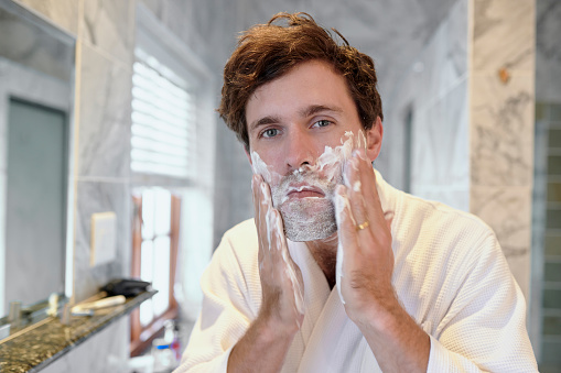Man, face and shaving cream in bathroom for skincare, grooming or personal hygiene at home. Male applying cosmetics, shave creme or product for facial hair removal, wash or treatment for clean skin