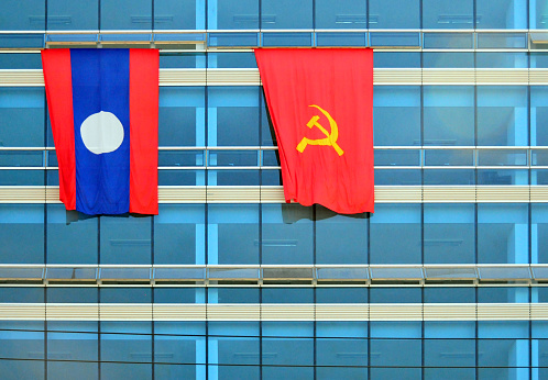 Vientiane, Laos: flags of the Lao People's Democratic Republic and the Lao People's Revolutionary Party (LPRP), the communist ruling party over a modern glass façade. The LPRP is the founding organization and single legal political party of the Lao People's Democratic Republic. The party's monopoly on government is enshrined in the Constitution, it administers a unitary state with centralised control over the economy and military.