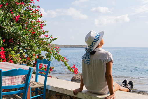 Woman sitting with typically Greek hat next to tavern and flowering plant with the Aegean Sea in the background on a sunny summer day