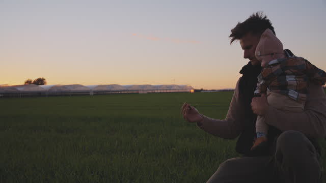 SLO MO Loving Male Farmer Showing Grass to Baby while Crouching on Agricultural Field during Sunset