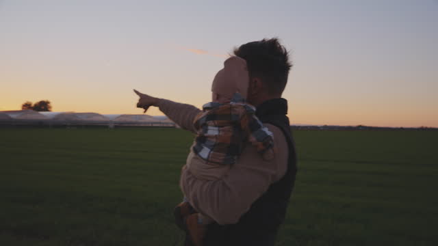 SLO MO Male Farmer Showing Agricultural Field and Greenhouse to Baby while Carrying him against Clear Sky during Sunset