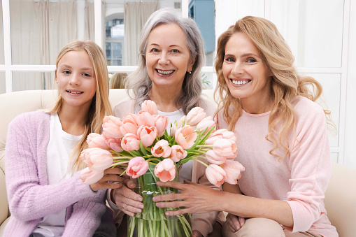 Mother and daughter giving grandmother bouquet of spring flowers, celebrate her birthday and taking photo for memory, sitting on couch