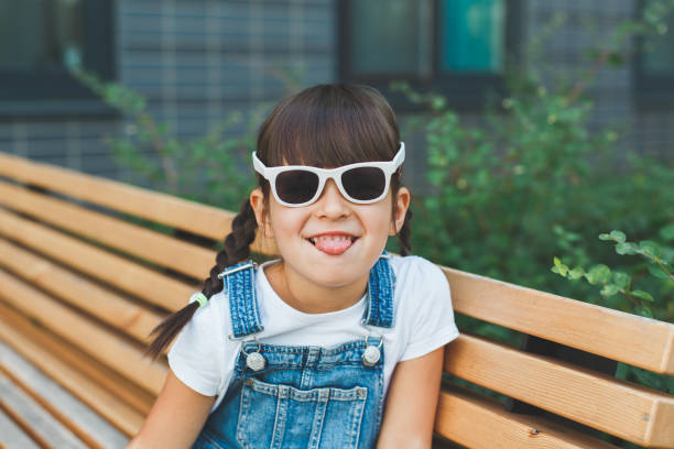 4 살짜리 귀여운 소녀는 여름에 선글라스를 착용하고 카메라를 보면서 얼굴을 찡그리며 벤치에 앉아 애지중지와 어린 시절의 개념을 보여줍니다 - cute 4 5 years little girls party 뉴스 사진 이미지