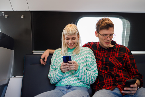 Portrait of young commuters sitting in metro train and using their phones. Travelers are using public transportation and commuting by subway. A happy couple sitting in metro train and typing on phones