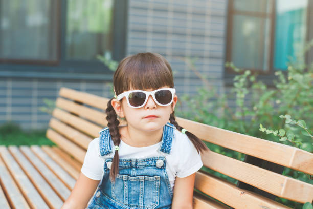 маленькая милая девочка 4 лет сидит на скамейке летом в солнцезащитных очках и смотрит в камеру, концепция баловства и детства - cute 4 5 years little girls party стоковые фото и изображения