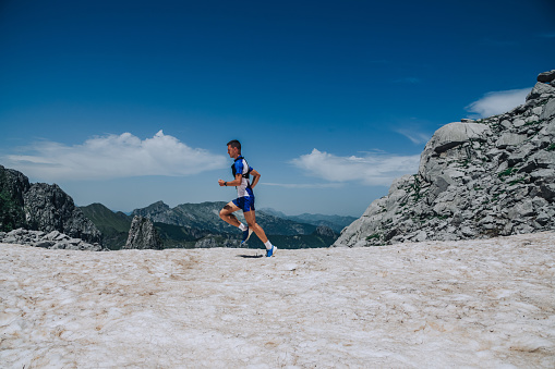An extreme sportsman is running fast, track running and skyrunning in mountains. A fit skyrunner is exercising and doing cardio workouts in wild nature in mountains. In background is cloudy blue sky.