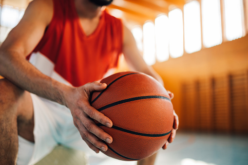 Close up of professional basketball player sitting on court with a ball in his hands. Cropped picture of an unrecognizable basketball player sitting on court during his training and holding a ball.